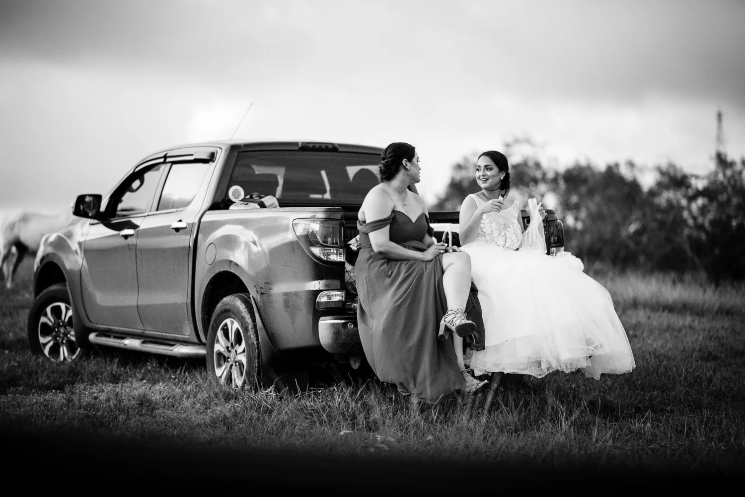 Two women sitting on the back of a pickup truck in an outdoor setting, one in a strapless dress and the other in a wedding gown, engaged in conversation, with trees and an open field in the background.