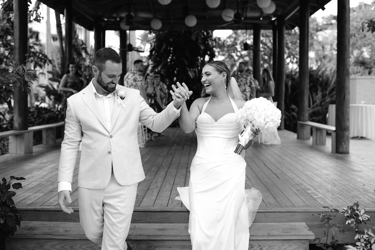 A bride and groom walk hand in hand, smiling happily, during their wedding ceremony on a wooden platform outdoors. The bride wears a white gown and holds a bouquet of white flowers, while the groom wears a light-colored suit with a boutonnière.