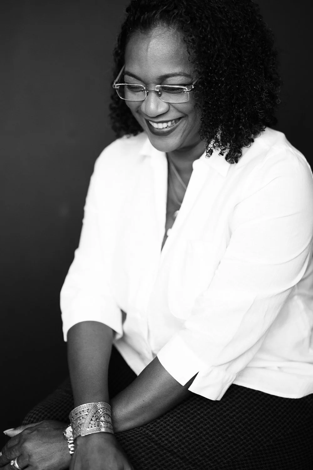 A smiling African American woman with curly hair and glasses, wearing a white blouse, sitting with her hands resting on her knees against a dark background.