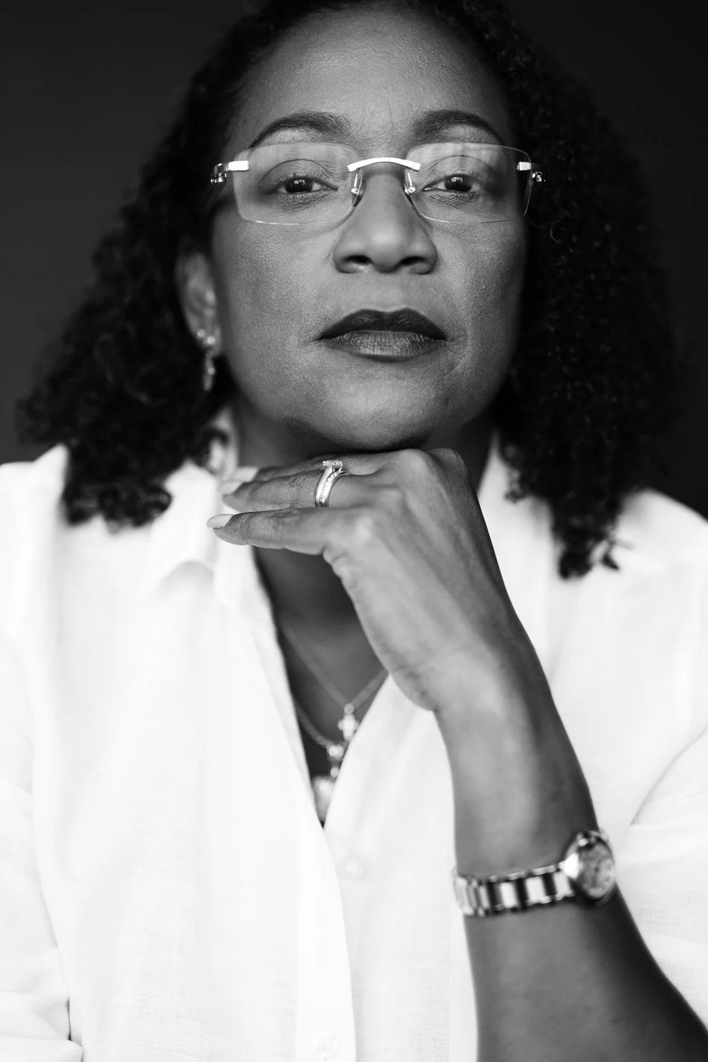 Black-and-white portrait of a confident African American woman with glasses, wearing a white shirt, sporting earrings and a wristwatch, resting her chin on her hand.