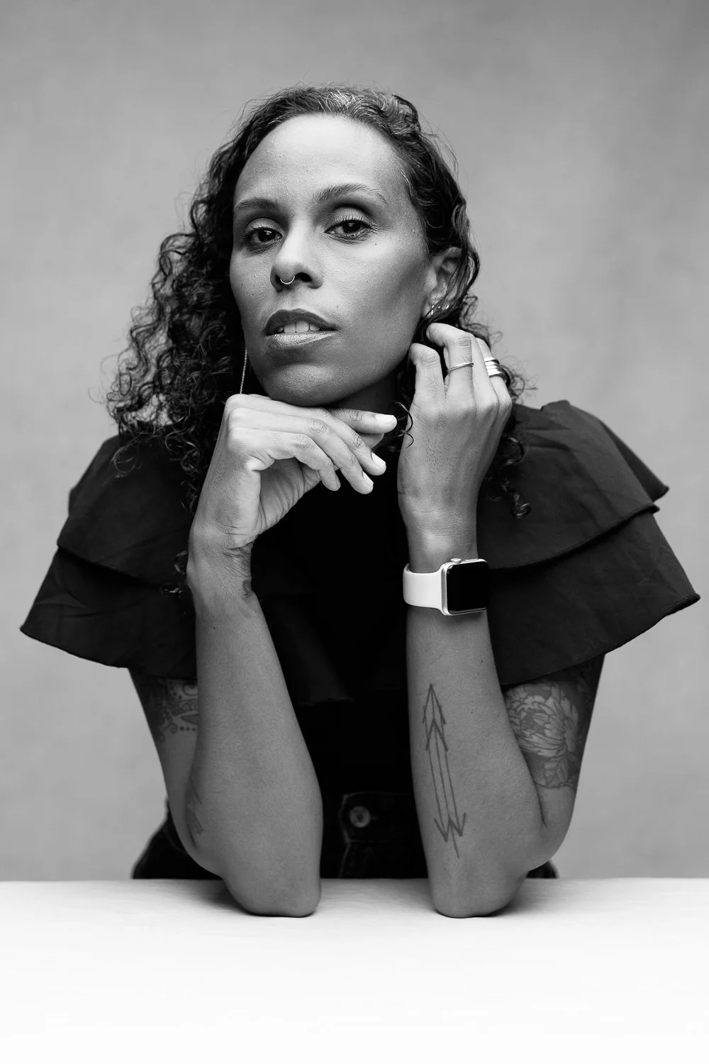A black and white portrait of a woman with curly hair, wearing a black top, with tattoos on her arms, rings, and a smartwatch, resting her chin on her hands against a plain background.