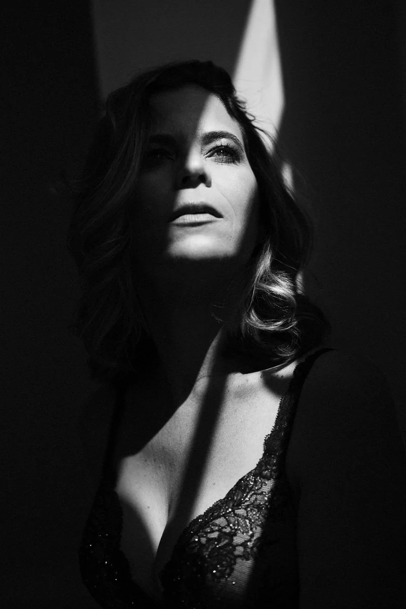 A black and white portrait of a woman with styled hair, wearing a lace top, illuminated by dramatic lighting.