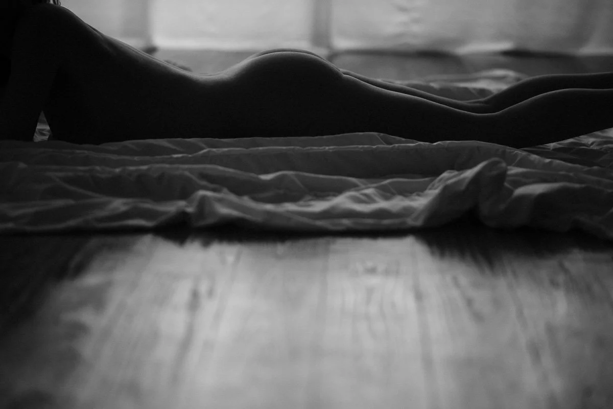 A black and white photo of a person lying on their stomach on a bed, with their back facing the camera, on a wooden floor with sheets or blankets crumpled nearby.