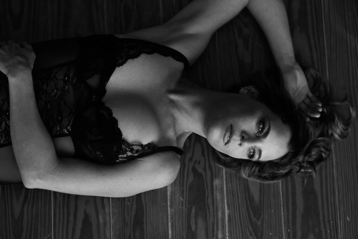 A woman with long wavy hair lying on a wooden floor, wearing a black lace dress, looking at the camera.