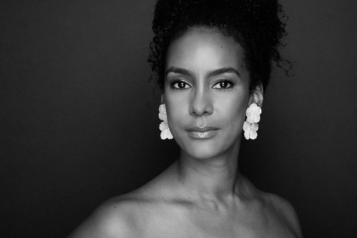 Black and white portrait of a woman with curly hair, wearing floral earrings, looking directly at the camera against a dark background.