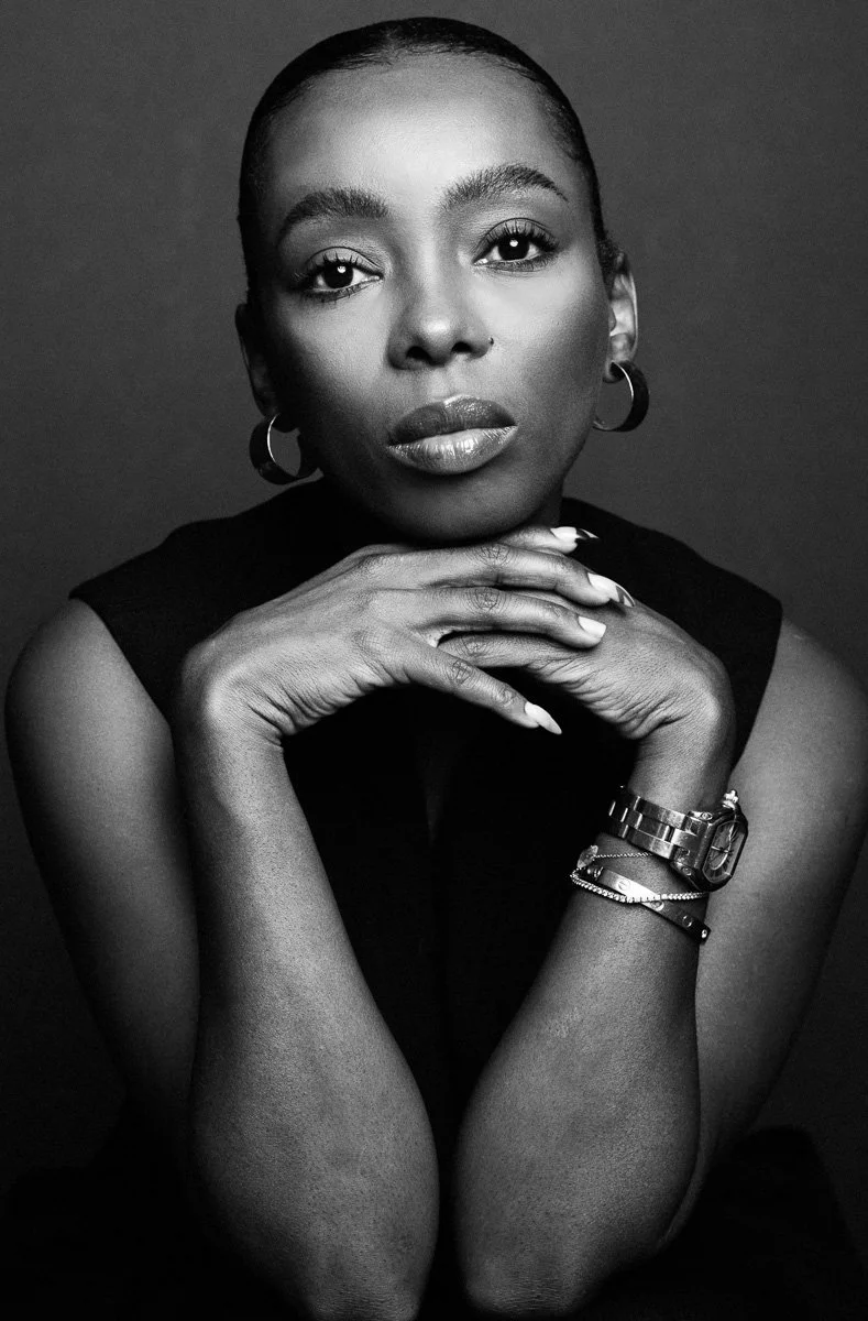 Black and white portrait of a woman with short hair, wearing hoop earrings and bracelets, looking directly at the camera with her hands gently resting under her chin.