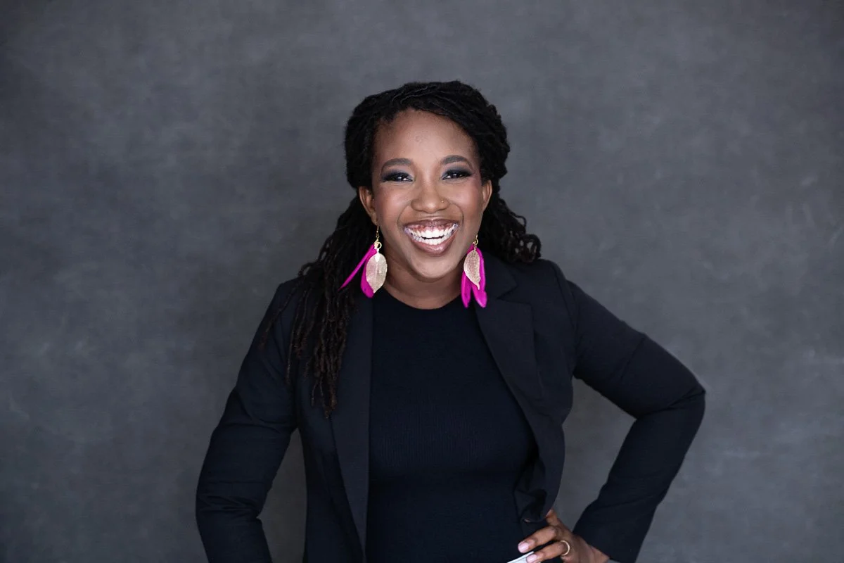 A woman with dark skin and long dreadlocks wearing a black blazer and top, smiling and posing against a gray background, with colorful earrings.