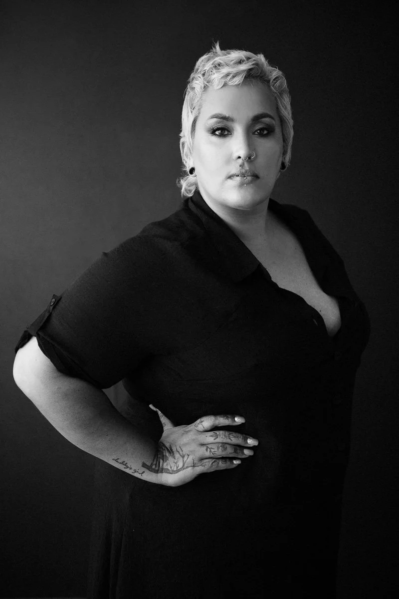 Black-and-white portrait of a woman with short, curly blonde hair, piercing eyes, multiple facial piercings, tattoos on her hands, wearing a black shirt, posing with one hand on her hip against a plain dark background.