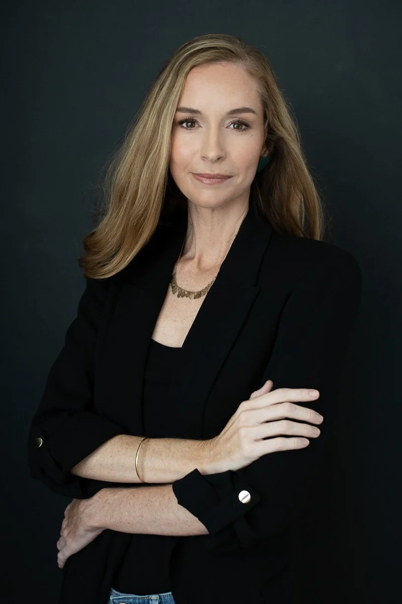 A woman with long blonde hair, wearing a black blazer over a black top, with a gold necklace, posing with her arms crossed against a dark background.