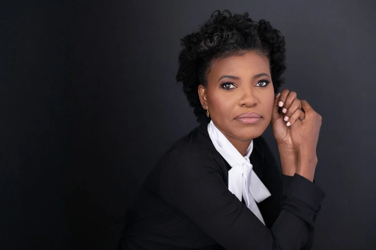 Portrait of a woman with natural curly hair, wearing a black top with a white bow tie, posed against a dark background.