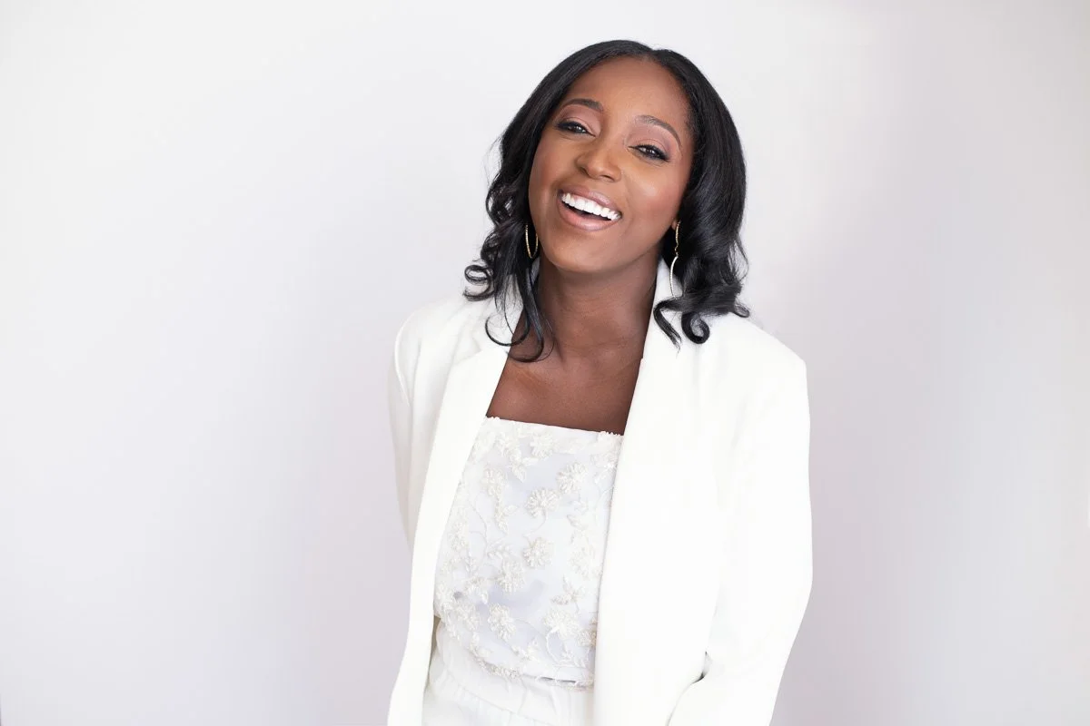 A woman with black wavy hair, wearing a white blazer and a white embroidered top, smiling with one eye closed, standing against a plain light background.
