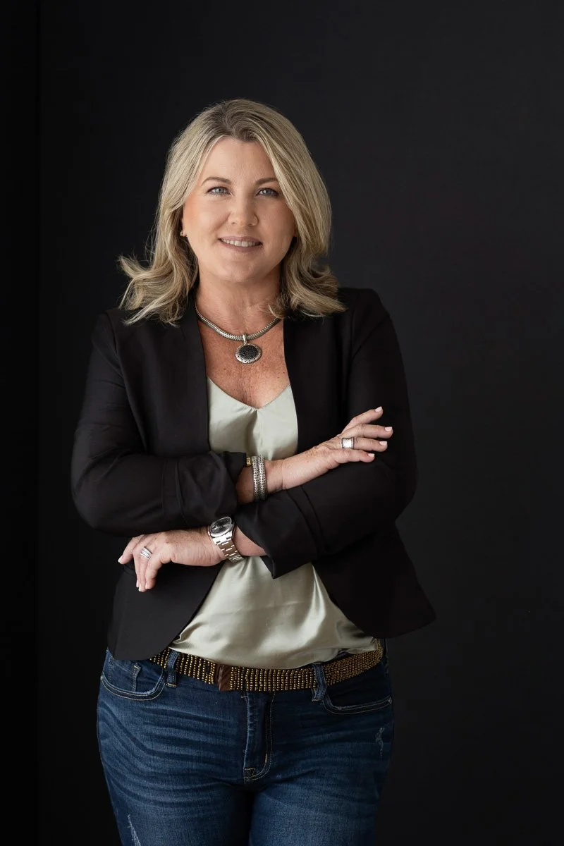 A woman with blonde hair, smiling, wearing a black blazer, a cream-colored blouse, and jeans, standing against a dark background.