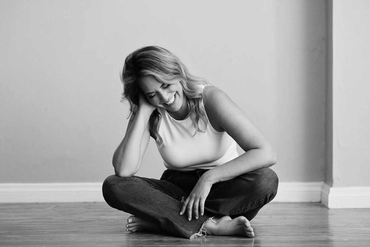 A woman sitting cross-legged on a wooden floor, laughing with her head tilted and hand resting on her knee, wearing a sleeveless top and distressed jeans.