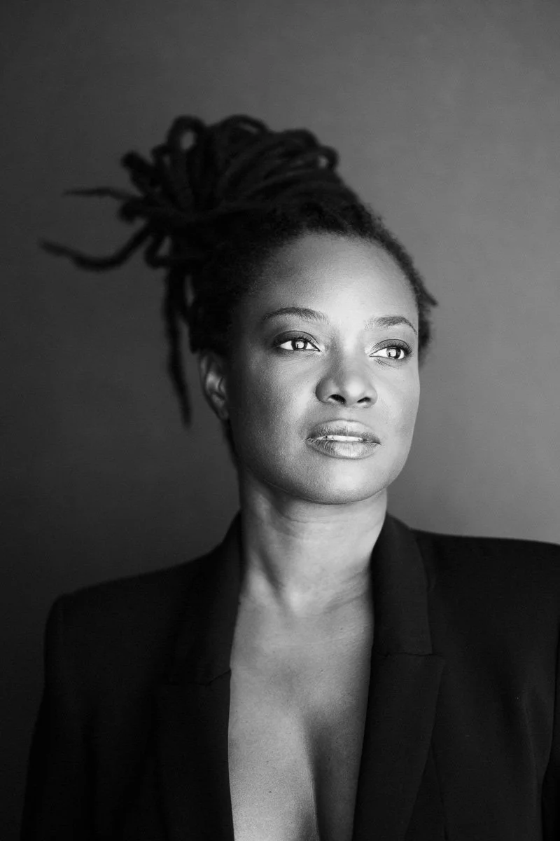 A black and white portrait of a woman with dreadlocks, wearing a blazer, looking slightly to the right.