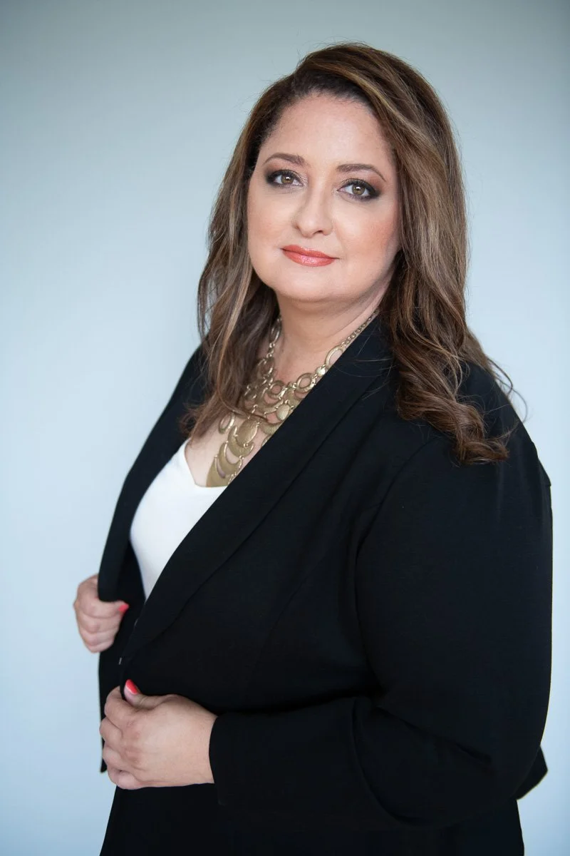 A woman with long brown hair, wearing a black blazer over a white top and a gold layered necklace, posing in front of a plain light background.