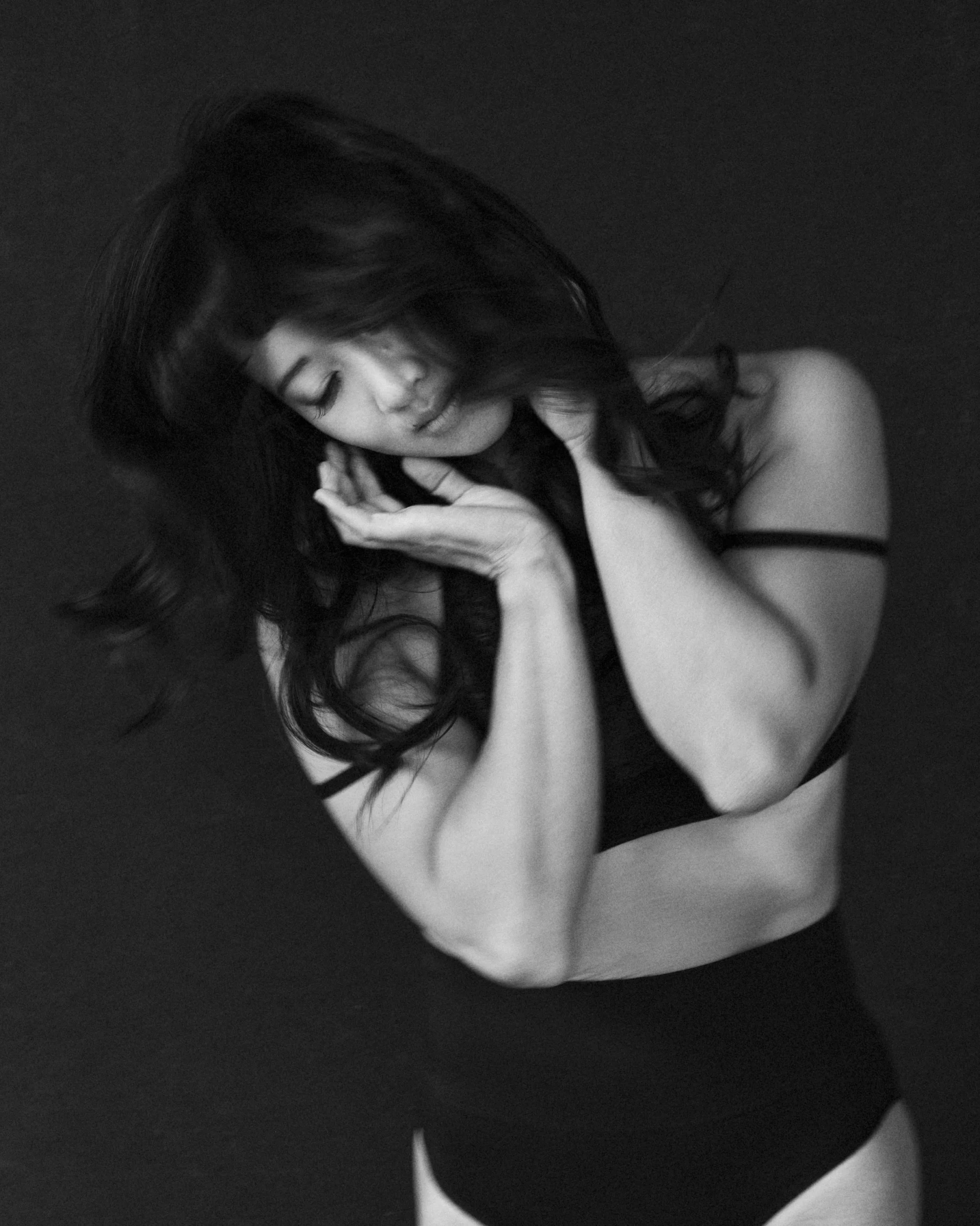 Black and white photo of a woman with long hair, eyes closed, touching her face and neck, wearing a black sleeveless top.