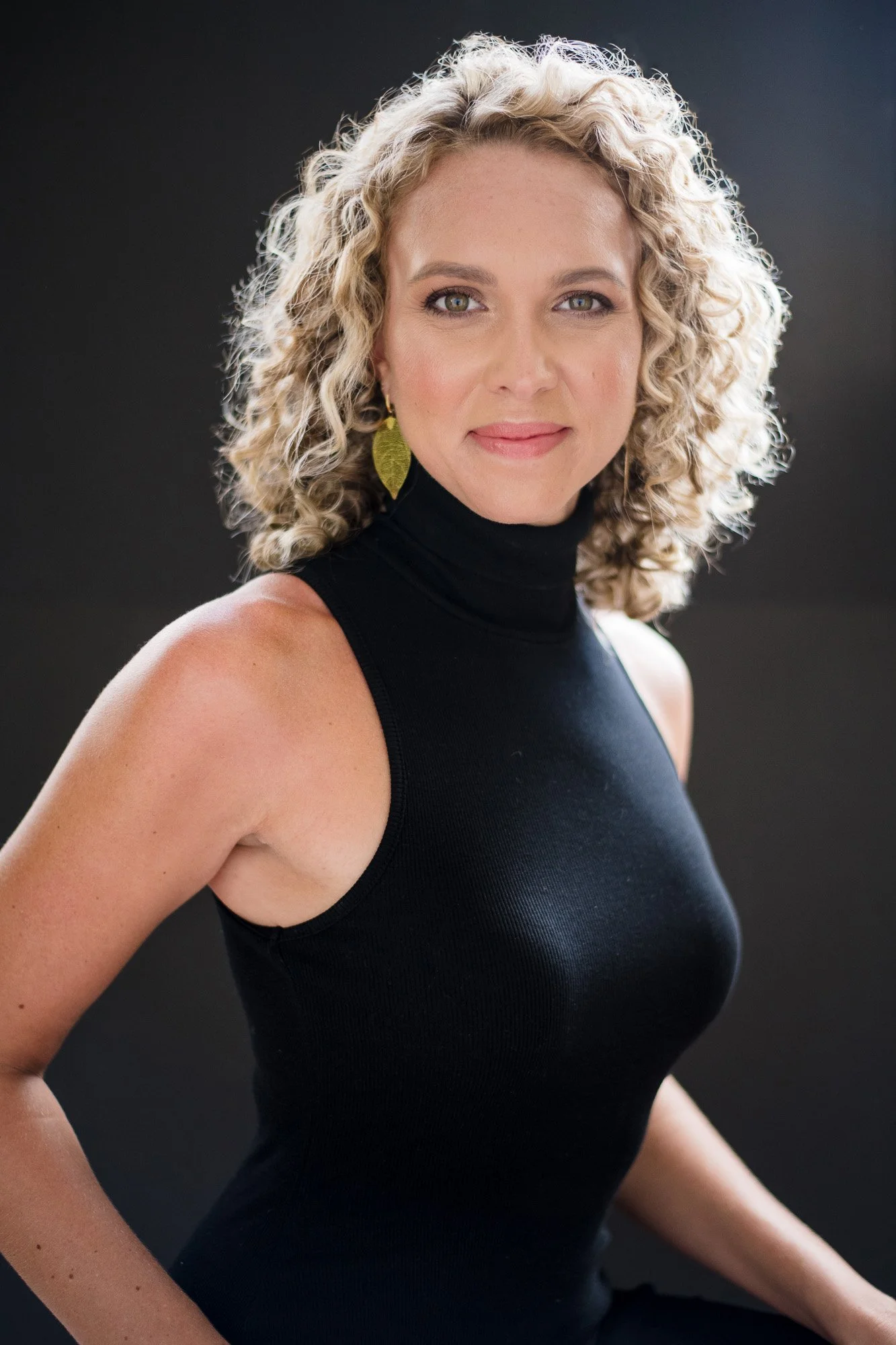 A woman with curly blonde hair wearing a sleeveless black turtleneck and gold leaf earrings, posing against a black background.