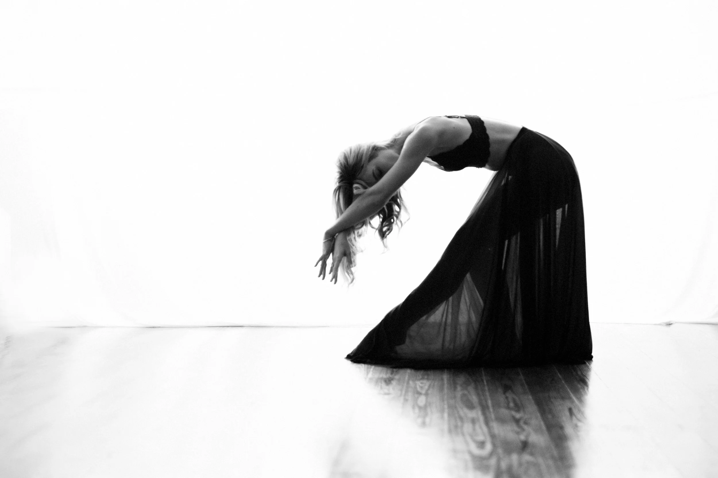A woman in a black dress performing a dance pose on a wooden floor against a plain white background, with her upper body bent forward and her arms extended downward.