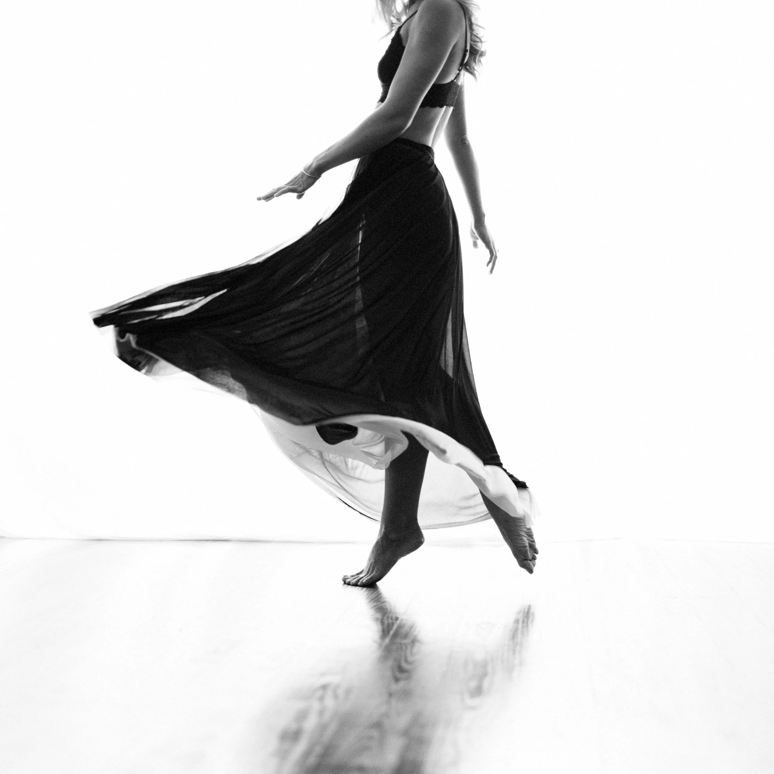 Black and white photo of a woman in mid-dance, wearing a flowing long skirt and a sleeveless top, with her feet on the ground and one leg slightly bent.