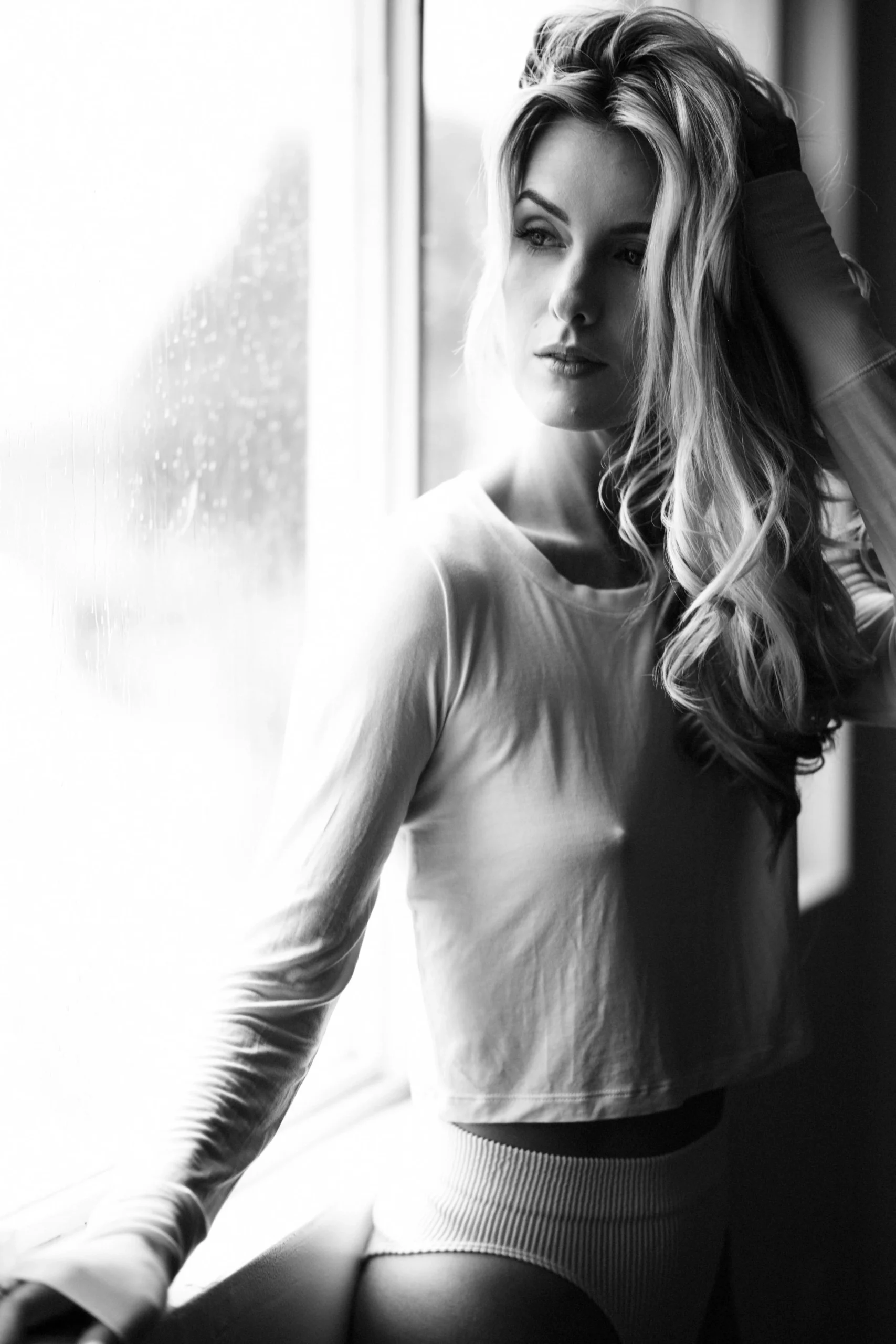 Black and white photo of a woman with long, wavy hair standing by a window, looking outside with a thoughtful expression.