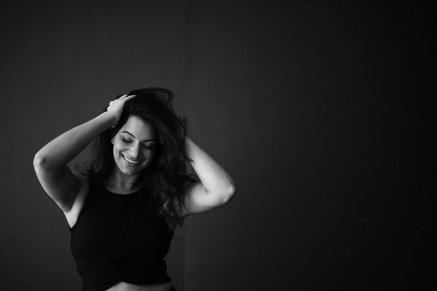 A black and white photo of a smiling woman in a sleeveless black top, with her hands in her hair, against a dark background.