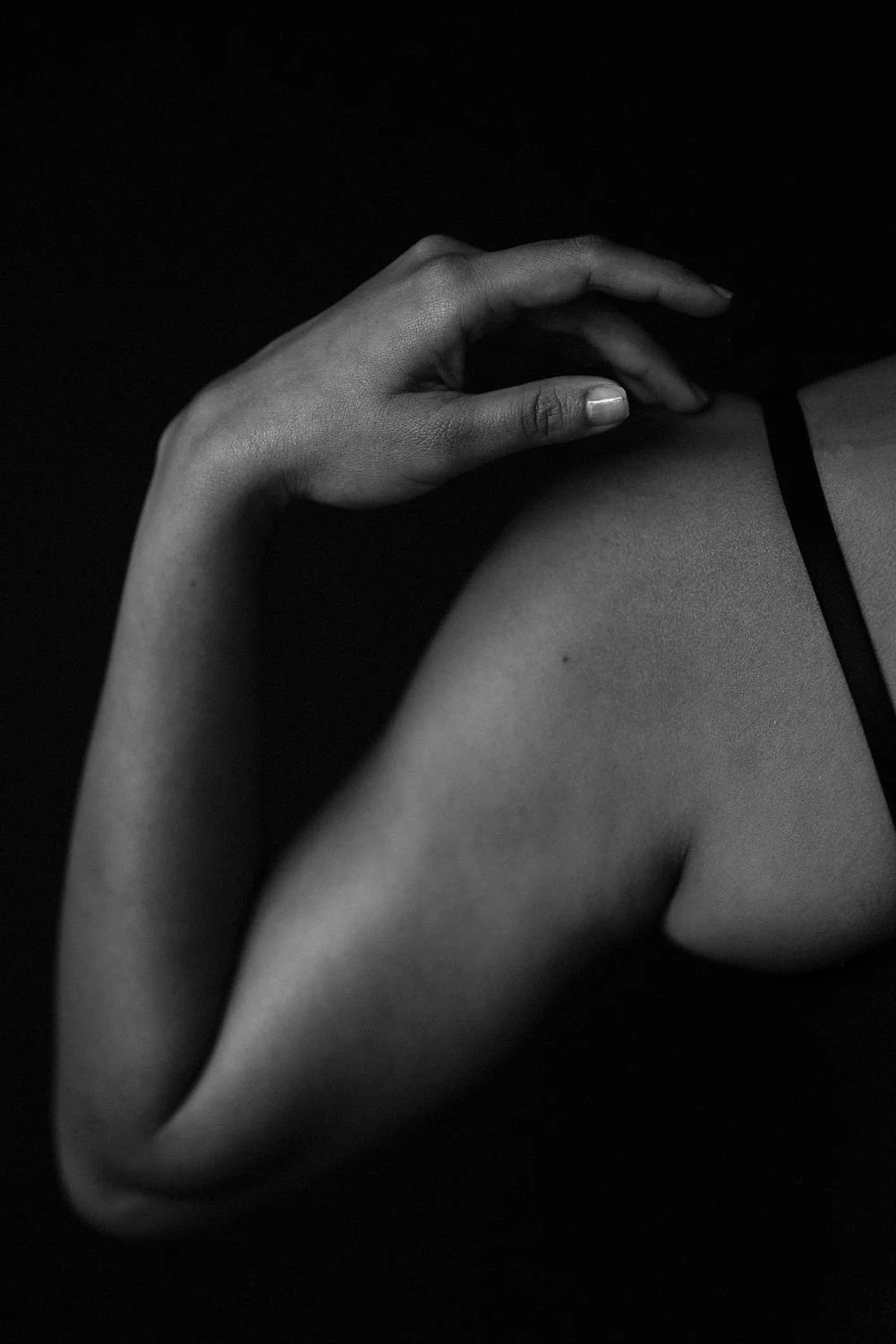 A close-up black and white photo of a person's hand resting on their shoulder, with the person's shoulder and part of their upper arm visible, wearing a thin strap top.