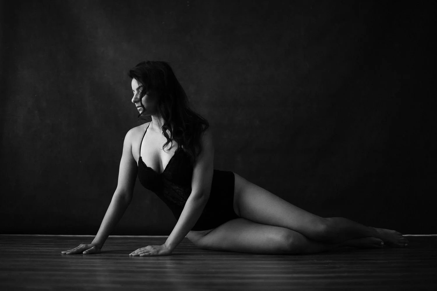 A woman in black lingerie positioned on a wooden floor, her body turned to the side, with a dark background.