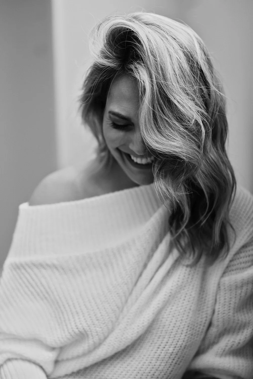 A woman with wavy hair smiling and looking down, wearing an off-shoulder sweater in a black-and-white photo.