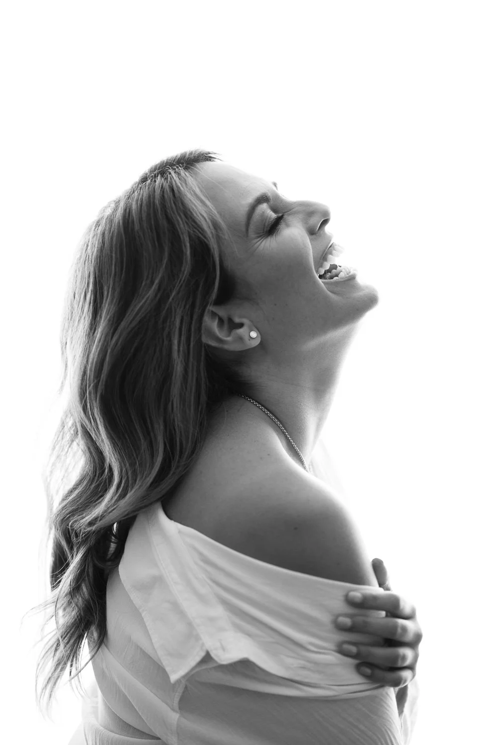 A woman with long wavy hair and earrings, smiling and laughing with her eyes closed, wearing an off-shoulder top, with her hand resting on her shoulder, black and white photo.