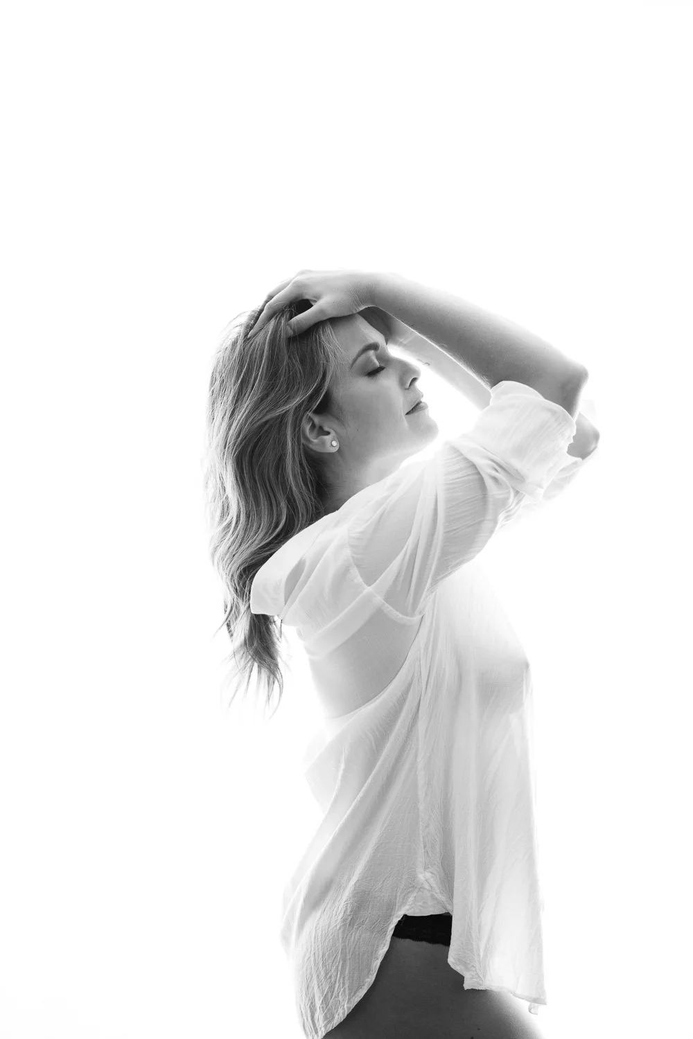 Black and white portrait of a woman with shoulder-length hair, eyes closed, smiling, wearing a loose white blouse, with one hand on her head.