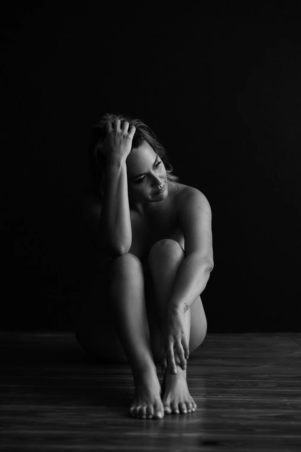 Black and white photograph of a woman sitting on the floor with her knees drawn up, her arms wrapped around her legs, and her hand resting on her head. She has short hair and appears to be deep in thought against a dark background.