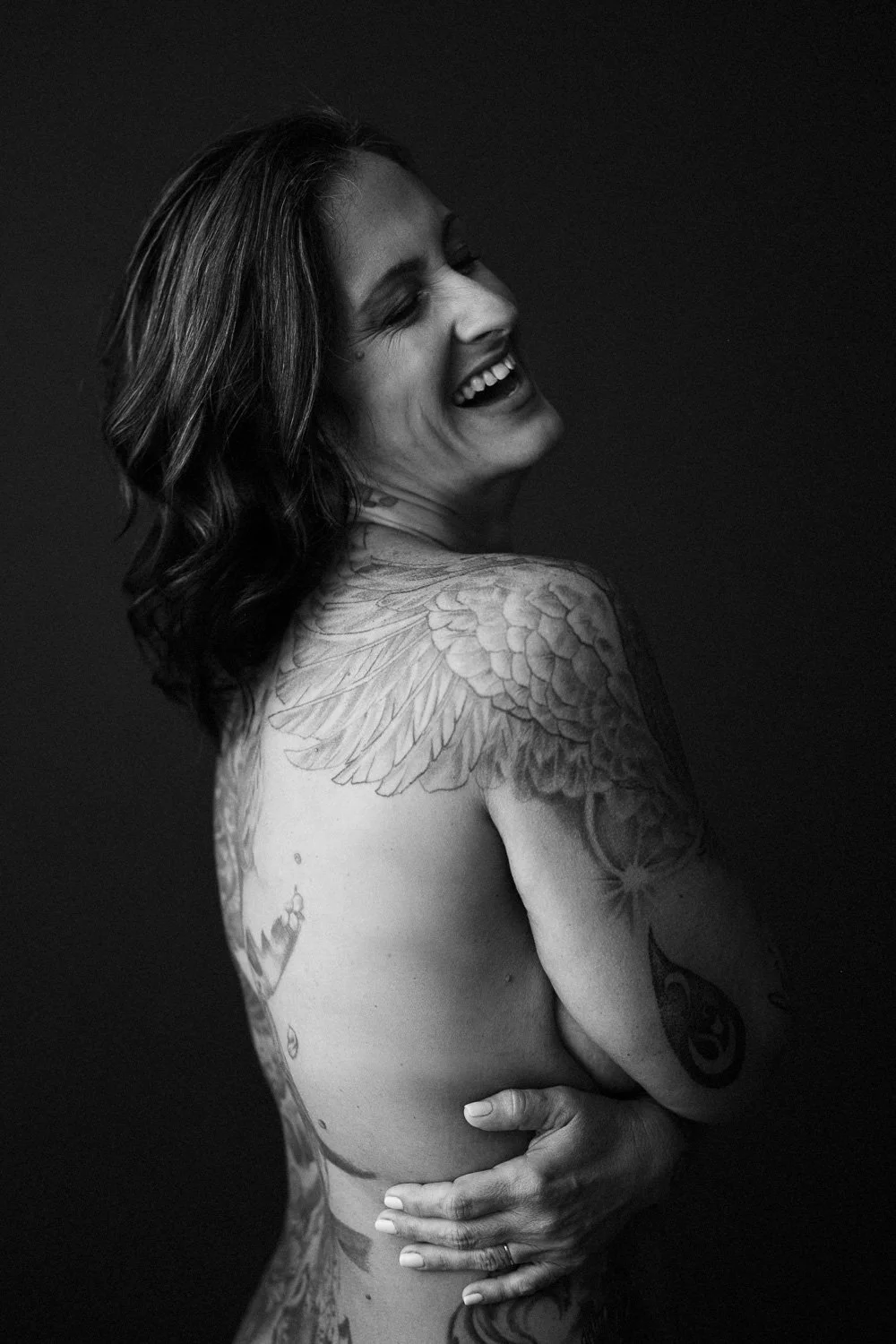 A black-and-white photo of a woman with shoulder-length wavy hair, smiling and looking to her left, with tattoos of wings and other designs on her shoulder, arm, and side.