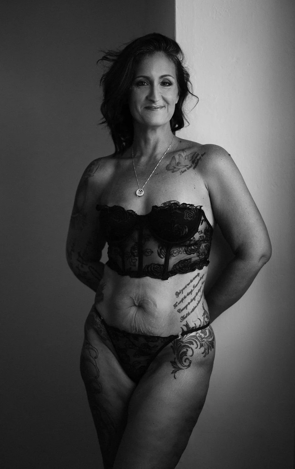 A woman with tattoos, wearing black lace lingerie, standing against a plain wall, smiling slightly.