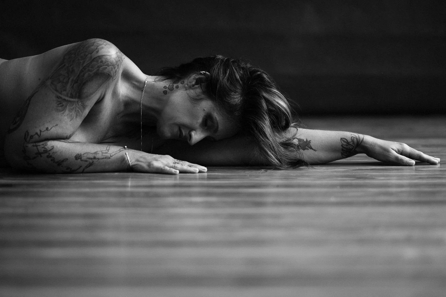 Black and white photo of a woman lying on the floor, with her head resting on her arm, looking at the camera. She has tattoos on her arm and neck, and her hair is slightly tousled.