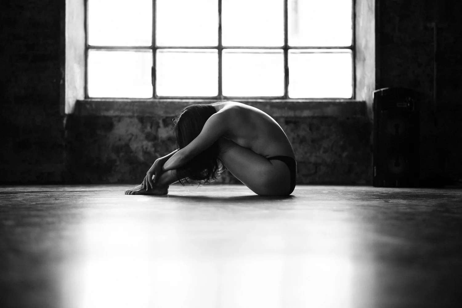 A person in black underwear sitting on the floor with knees bent and arms wrapped around legs, head resting on knees, in a dimly lit room with large window and exposed brick wall.