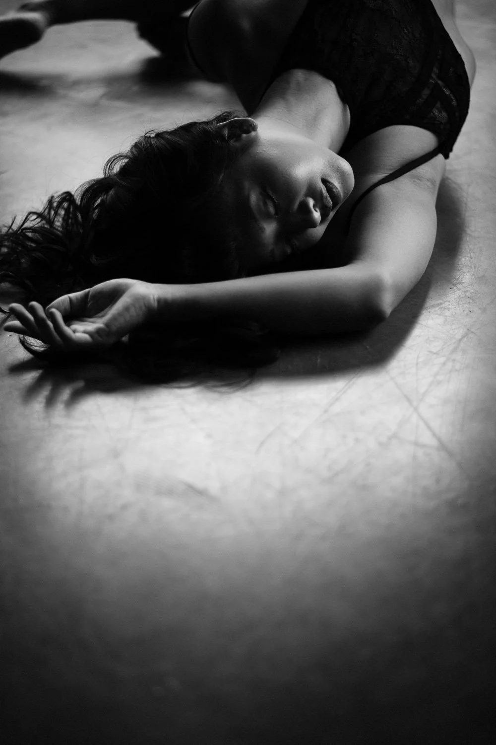 A black-and-white photograph of a woman lying on the floor with her eyes closed and arm stretched above her head.