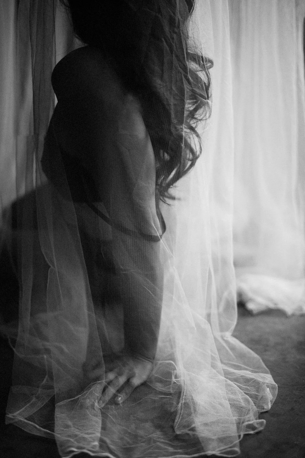 A woman with long curly hair is seen kneeling behind a sheer curtain, wearing lingerie, with her face partially obscured.