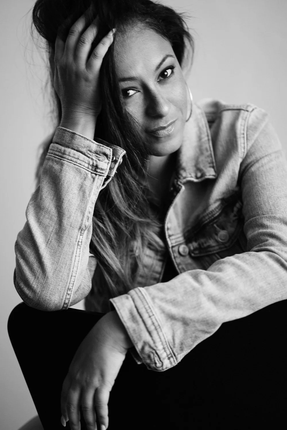 Black and white portrait of a woman with long hair, wearing a denim jacket, looking at the camera with one hand on her head, sitting on a chair.