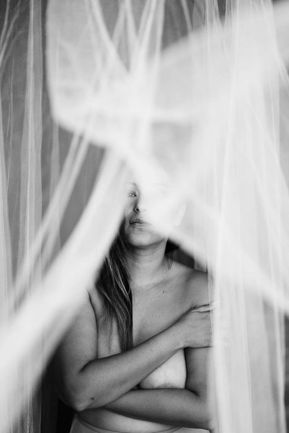 Black and white photo of a woman partially covered by sheer fabric, with one arm crossed over her chest and her face partially obscured by the fabric.