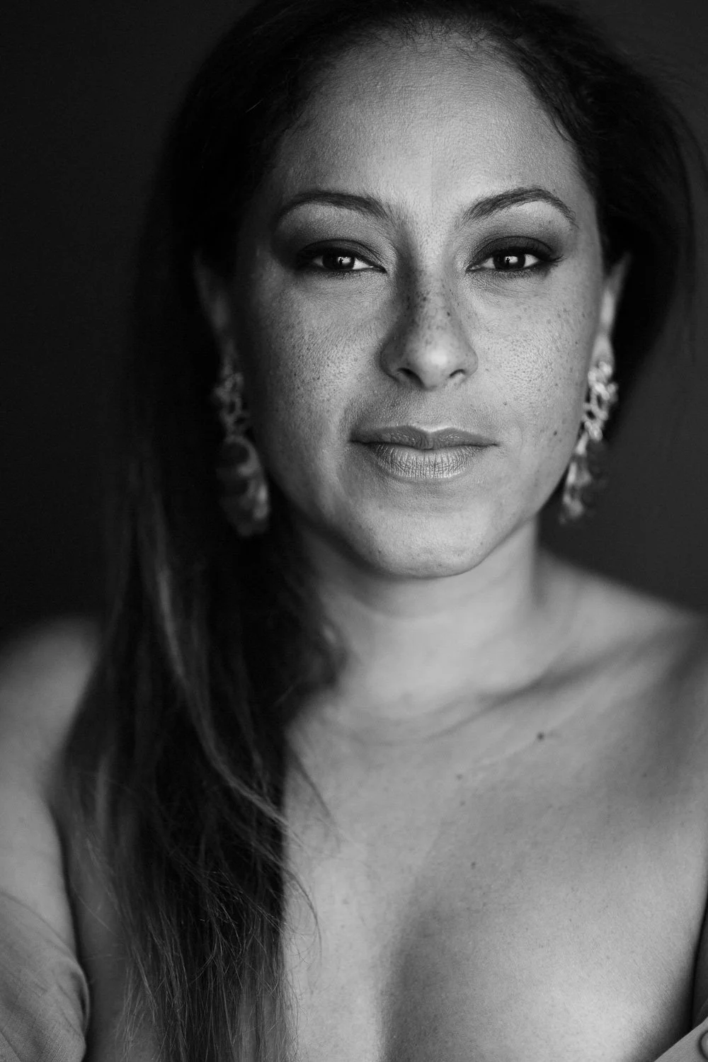 Close-up black and white portrait of a woman with dark hair, expressive eyes, and earrings, looking directly at the camera.
