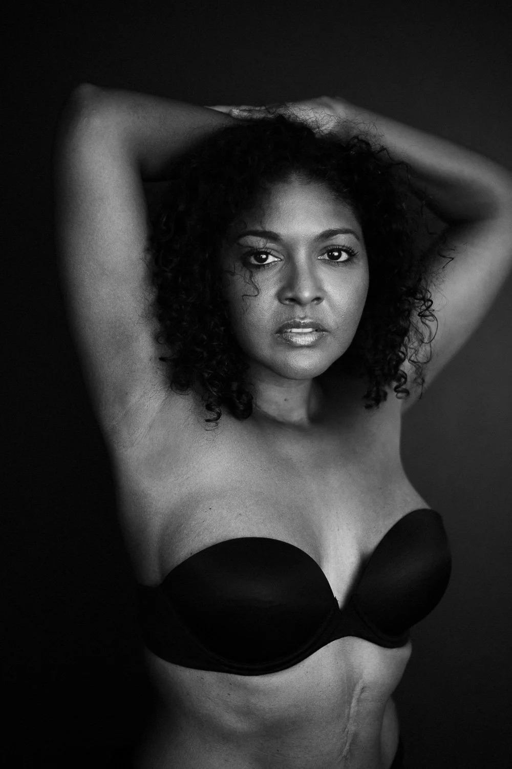 Black and white portrait of a woman with curly hair, wearing a strapless top, posing with arms raised and hands behind her head against a dark background.
