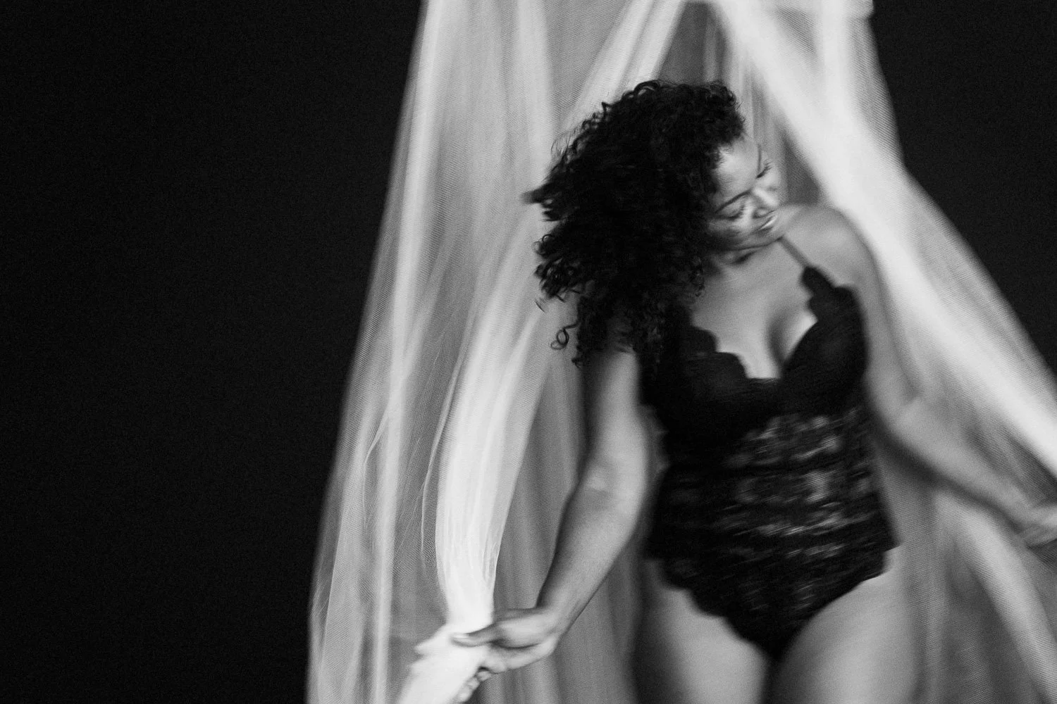 Black-and-white photo of a woman with curly hair wearing black lingerie, holding a piece of sheer fabric above her head.