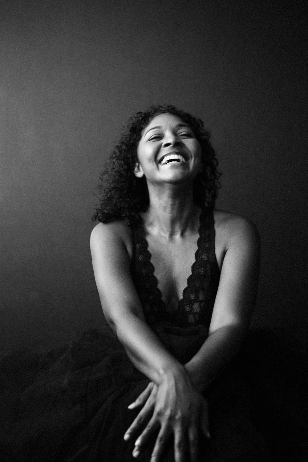 Black and white photo of a woman with curly hair, smiling joyfully, wearing a sleeveless dress with lace details, sitting against a plain wall.