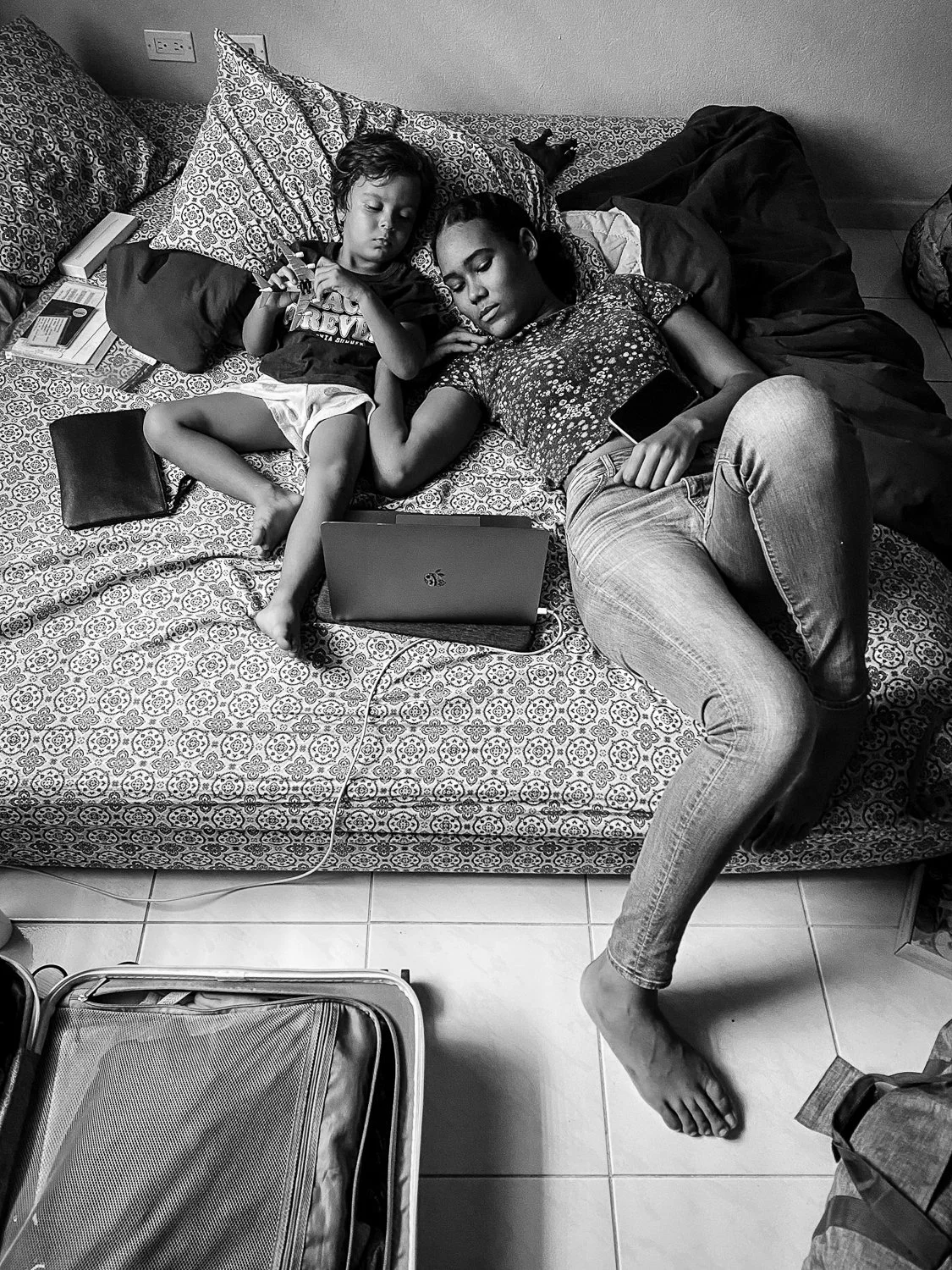 A woman and a young boy are lying on a bed, sleeping. The woman is holding a smartphone, and there is a laptop, a closed book, and some other objects on the bed. The woman is wearing jeans and a floral top, while the boy is in a T-shirt and shorts. T