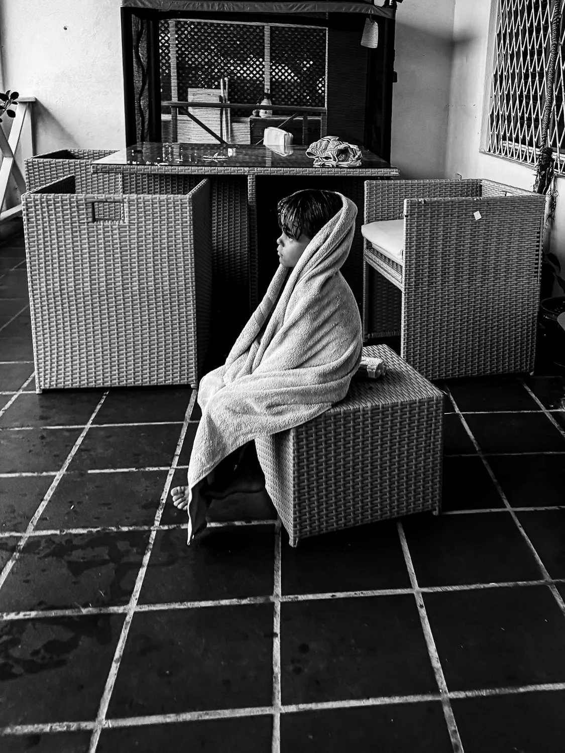 A young boy sitting indoors on a wicker chair, wrapped in a towel, with wet hair, in a space with tiled floors and outdoor furniture.