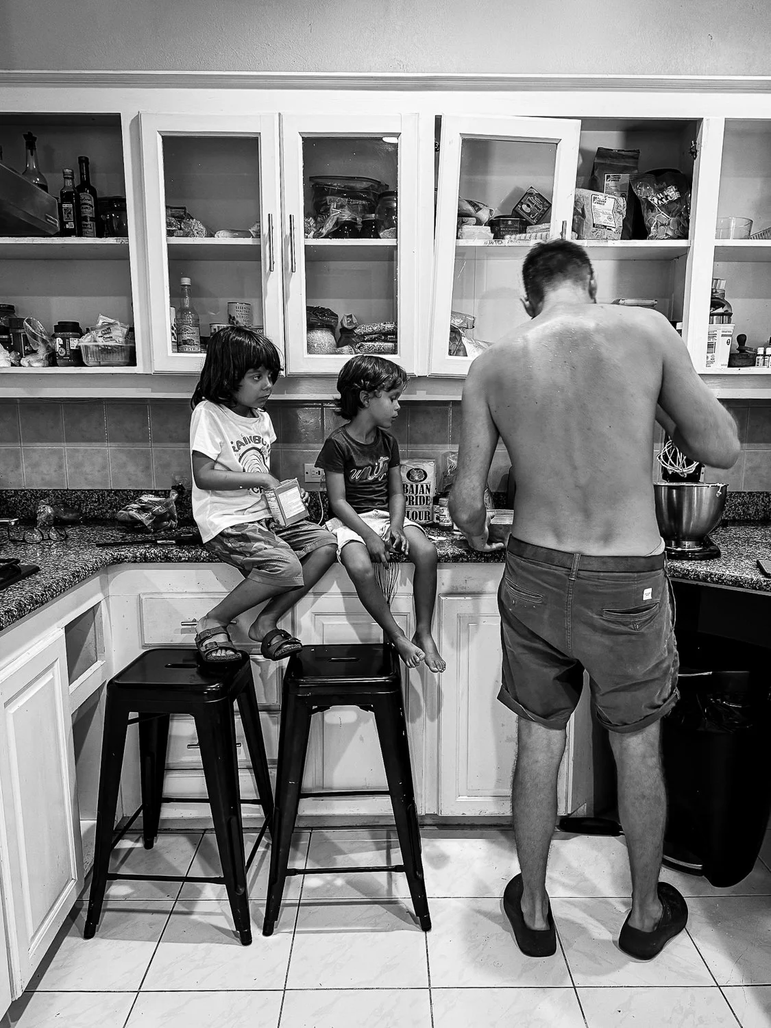 A man with bare back and shorts is cooking in the kitchen while two children sit on stools on the counter, one holding a box, in a black-and-white photo.