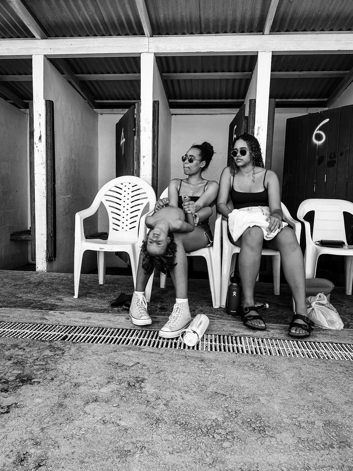 Three young women sitting on plastic chairs against a wall with changing rooms numbered 4, 5, and 6. One woman is upside down, draping her head over a chair, wearing sneakers, shorts, and sunglasses. The second woman is adjusting her sunglasses, wear