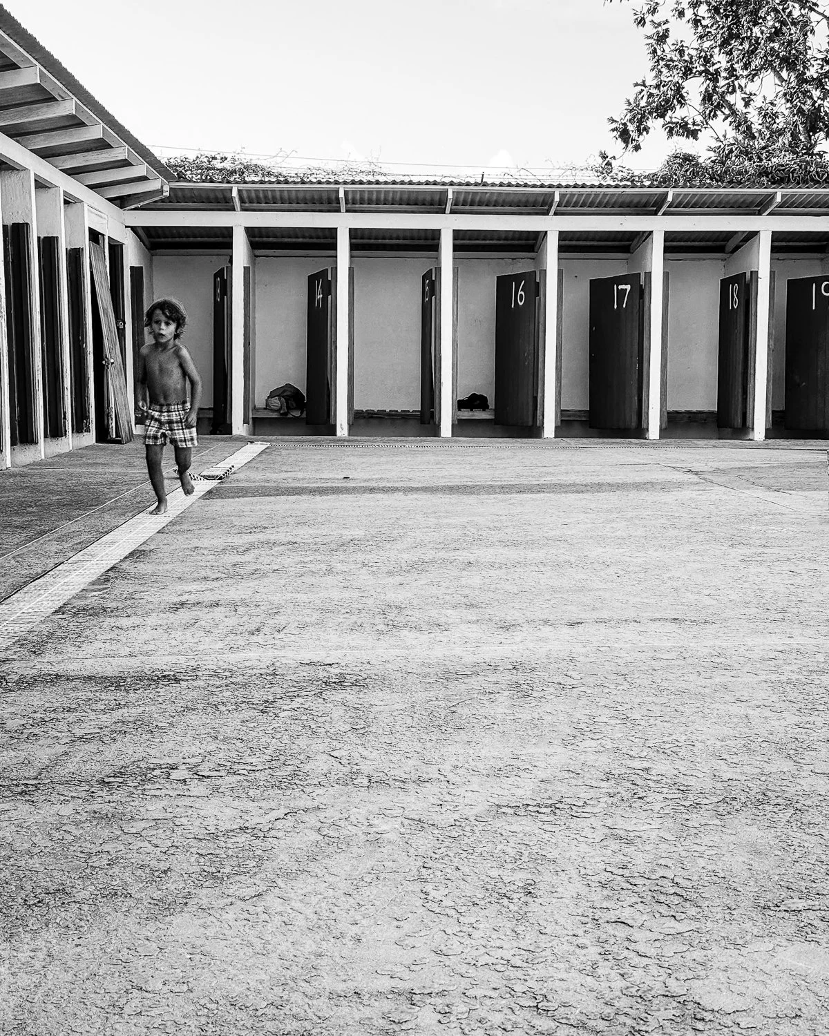 A young boy in plaid shorts walking barefoot in an outdoor shower area with numbered wooden changing stalls in the background.