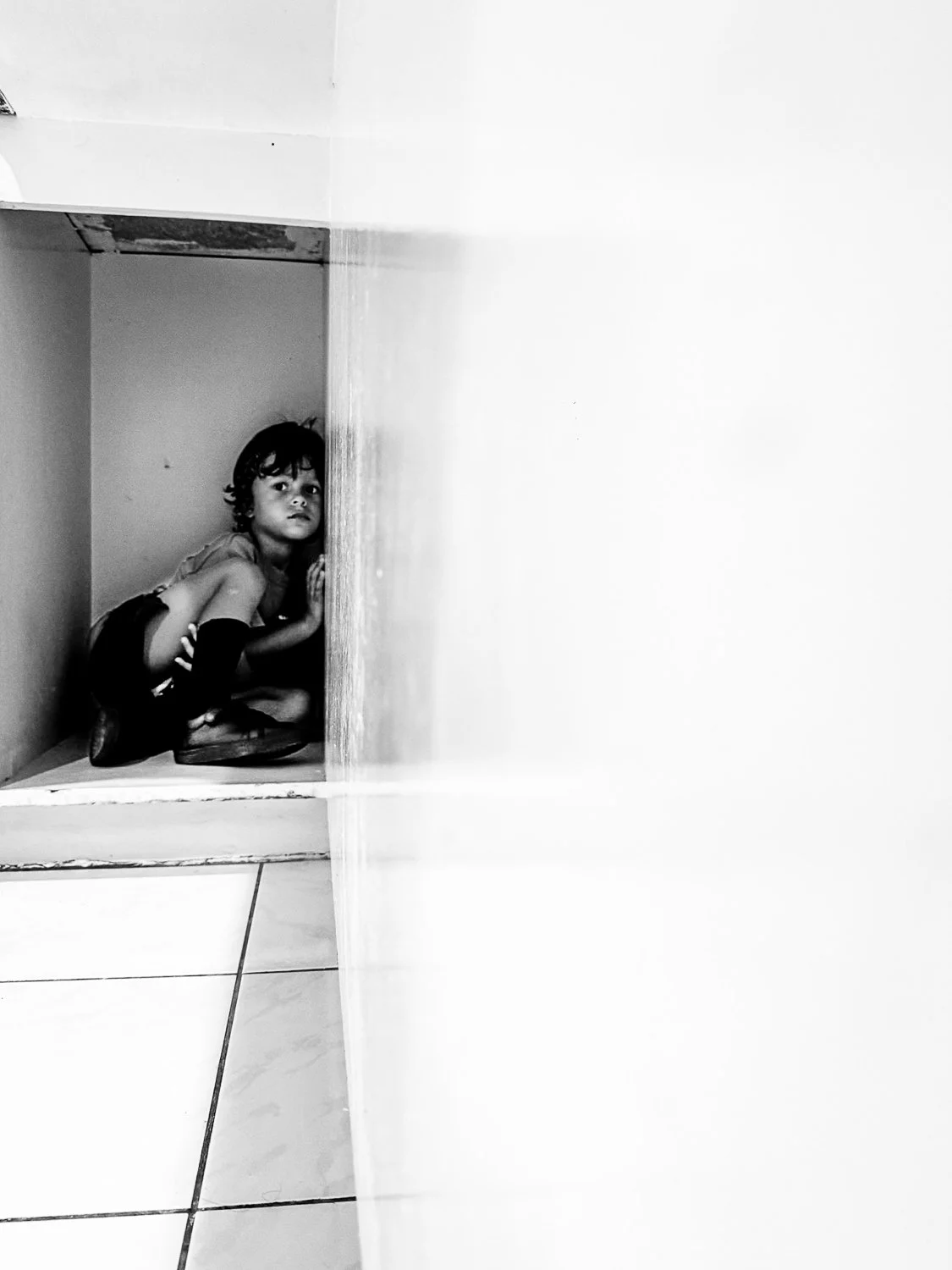 Child crouching in a small corner, looking around the corner in a black and white photo.