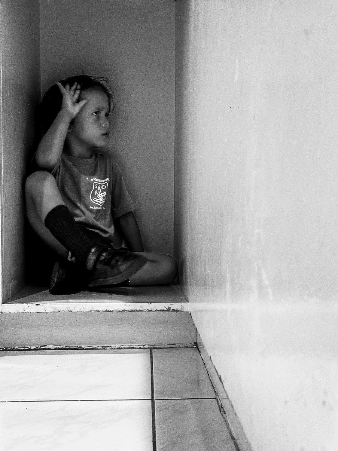 A young boy sitting in the corner of a small nook, with his legs crossed, looking thoughtfully into the distance, hand resting on his head.